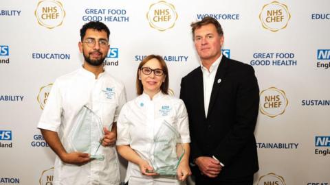 The image is of three people. On the left is a man wearing glasses with dark brown hair. In the middle is a woman wearing glasses with light brown hair. Both are smiling and holding glass trophies. On the right is a man in a black suit. 