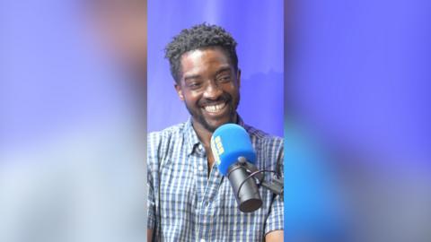 A man with short, dark hair smiles as he talks into a microphone