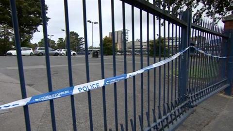 A line of police tape is stretched across metal gates which are painted blue. Behind them we can see parked cars and buildings in the distance