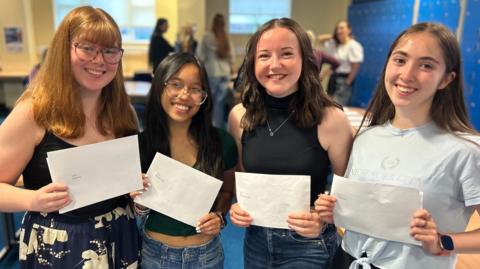 Four female A-level students in summer clothing smile as they hold up white envelopes. Two of the teenagers are wearing glasses.