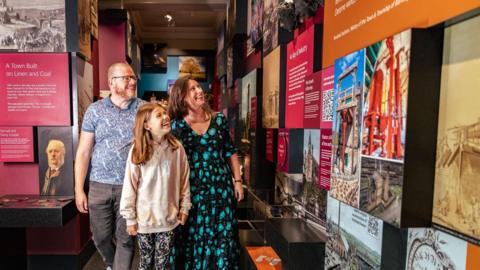 A girl, aged about 10, in a pink hoodie and leggings, walking through a museum corridor with her mum and dad behind her. Dad wears a blue paisley polo shirt and mum wears a black and turquoise patterned summer dress. The walls are covered with information and photos.