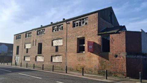 An exterior picture of the former Gleadells Wharf building. It has broken windows on the top floor, with the lower ones boarded up. There is a footpath to the side with black bollards and a road.