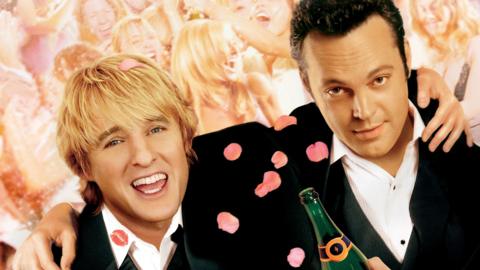 Two people in dark suits stand close together at a crowded wedding reception. Pink petals and lipstick marks are visible on the suits, with a blurred dancing crowd behind.