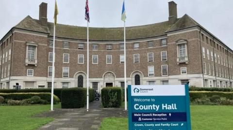 Somerset Council's County Hall pictured on a cloudy day. The town hall is in the background of the image. In the foreground is a sign which says 'Welcome to County Hall'. There are three flags poles outside of the building.