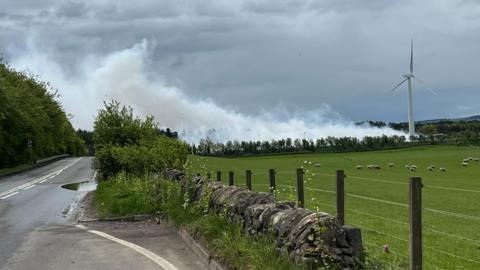 White smoke coming from an area behind a hedge and next to a wind turbine. There is a field containing sheep in the fireground and a road to the left of the picture. The snoke is blowing over the road. 