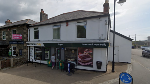 A Morrisons shop in a two-storey white painted building with a side entrance andlean-tp next to a road at the end of a line of shops.
