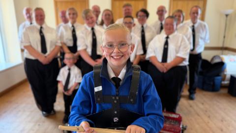 Evie wearing a blue band jacket and a white shirt and tie smiling at camera. She has a black drum strap on her and drum sticks. in her hand. Her band members are in the background wearing white shirts, a black tie and black trousers. The floor is wooden. They are standing in a room. 