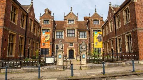 The image shows the outside of a museum, which has two banners on either side of the building: the one on the left is orange with a fossil on it, and the banner on the right is yellow with what appears to be a bird on it. 
There are black fences in front of the main entrance, with a gate in the middle of two pillars. The main black door can be seen in the background down a path.