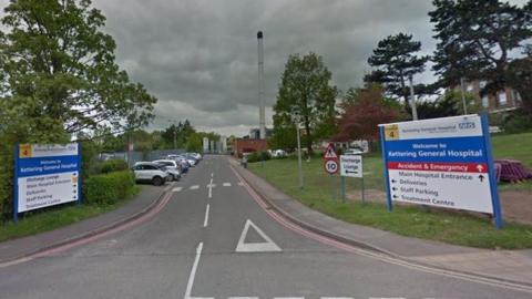 Entrance to Kettering General Hospital showing two-lane road heading past signs on either side, with black writing on a white background and a blue band towards the top on which Kettering General Hospital is written out in white lettering. There are cars parked in spaces on the left hand side of the road and trees to the left and right.