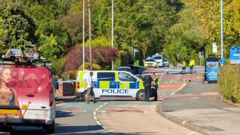 A police van parked across a road with officers seen around the scene and police tape across the road