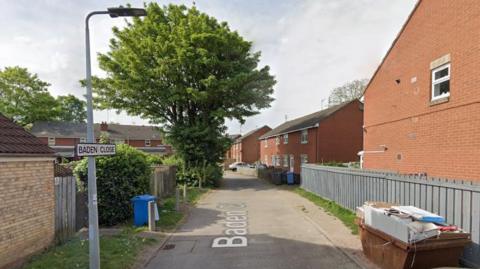 A tree-lined lane that leads to houses on Baden Close, there is a lamppost with the road name sign on and a skip filled with rubbish.