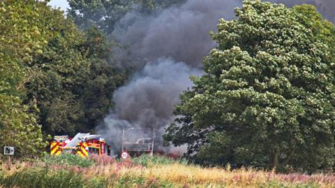 A fire-damaged bus in the distance with smoke going up in the air next to a fire engine surrounded by trees and a 50 speed sign