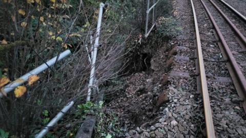 The side of a railway track which shows stones on the left crumbling down a bank. On the right is the bronze railway track. To the left of the fallen stones there are bushes.