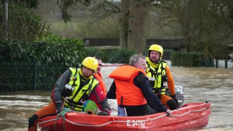 Two men wearing life jackets sit in a red rescue boat with two rescue workers on flooded waters