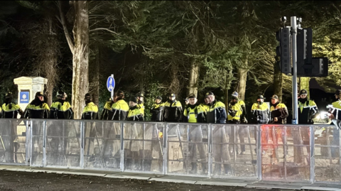 A row of a couple dozen men in hi-viz police jackets. It's dark and there are tall green trees behind them. A steel silver fence separates the police form the road. 