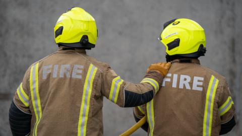 Two firefighters in uniform. One of them has his hand on the other.