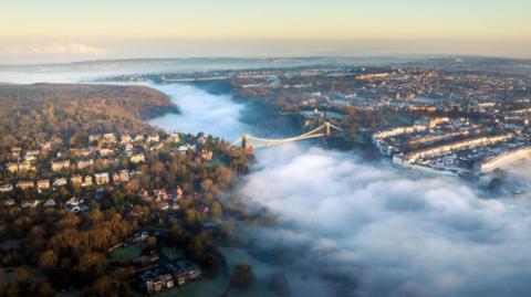 An aerial view of the Avon Gorge during sunrise. The sky is a pale blue with orange and pink stripes, and a low fog is rolling through the valley. The Clifton Suspension Bridge is stretching across the river above the mist.
