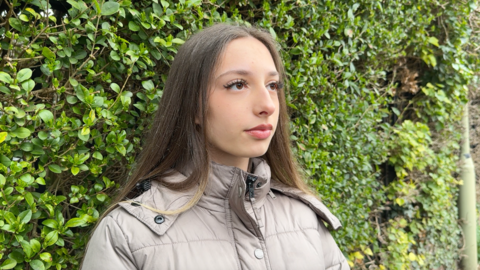 Image shows a student called Megan. She has long brown hair. The ends are blond. She is wearing a beige puffer coat which is zipped up. She has a neutral expression and is looking off to the right of the image towards the reporter, who is asking her questions. Behind her, the whole background is filled with green from a large green bush. 