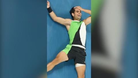 Carlos Alcaraz smiles on a blue floor wearing a green, black and white vest and shorts. He has one hand on his head.
