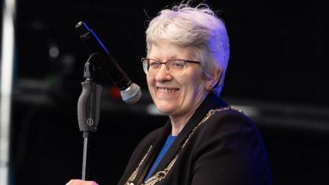 A woman with glasses and grey hair smiles in front of a microphone. She is wearing a black jacket with a blue T-shirt underneath and has a gold chain of office around her neck