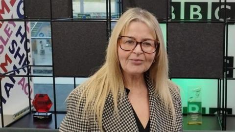 A woman with long blonde hair wearing a black and white check double-breasted jacket and brown-framed glasses.