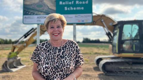 A woman with short blonde hair wears a leopard-print top and long necklace. She is smiling as she stands in a field in front of a large sign reading "relief road scheme". A large yellow excavator is parked behind the sign.