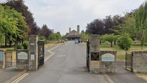 The entrance gates to Coney Hill Cemetery and Crematorium in Gloucester. Big stone posts with metal gates lead to a long driveway between green spaces with trees to the crematorium building.