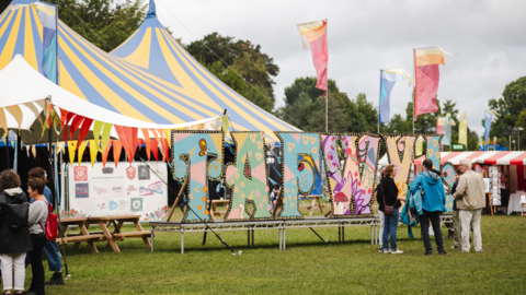 The Tafwyl sign in a field with tents in the background and people gathered chatting in groups