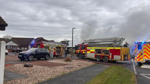 Fire engines and an ambulance gathered around a house with smoke billowing into the air
