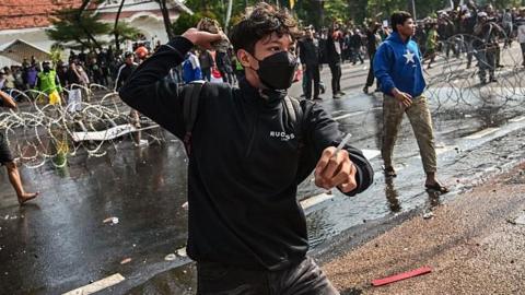 A young man, wearing a black face mask and a rucksack, appears to hold a rock back ready to throw on a busy road during demonstrations