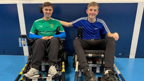 Two brothers sat in mechanical chairs inside. They're both smiling and the boy in the blue top has his arm round the other's shoulder. George, on the left, is in a green football shirt.