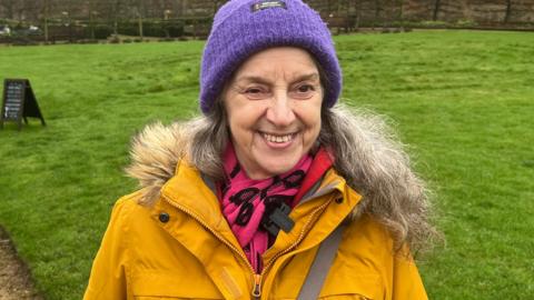 York's Babs Callaghan - who is pictured at Helmsley Walled Gardens - is one of the first people to be supported by the new "Natural Health Service. Babs is smiling, and wearing a yellow parka coat. She is wearing a lilac woolly hat and is standing in front of one of the garden's lawns.