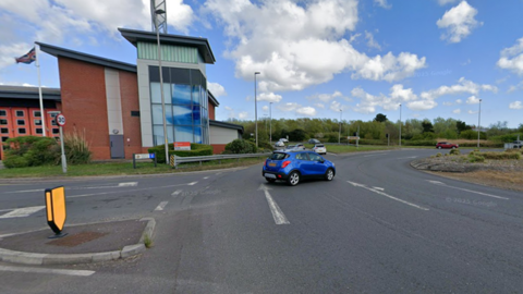 A Google Streetview image of the roundabout junction with a small blue car on the carriageway and a fire station building in the background