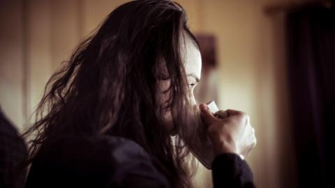 A stock photo of a woman with long brown hair which is partly over her face drinking from a mug. She is wearing a dark top. 