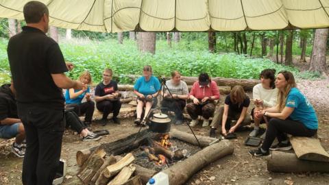 Young people sitting on logs around a camp fire as they listen to an instructor. They are sitting underneath an open canopy with trees and woodland in the background.