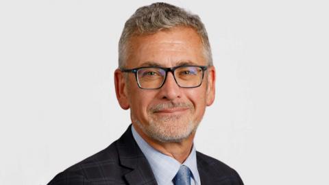 Prof Shane O'Neill is smiling in a university website picture. He has grey hair and beard and is wearing dark glasses, a dark jacket and grey shirt and tie