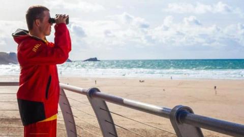 The photograph is taken from an RNLI vantage point across the beach. A volunteer stands to the left of the picture in a red waterproof suit. He is looking through binoculars acros the beach.
