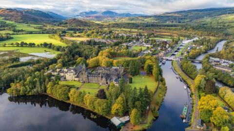 An aerial view over the picturesque village of Fort Augustus on the shores of Loch Ness. The village's abbey is in the foreground next to the loch and a section of the Caledonian Canal, with its locks. There are lots of trees dotted through the village and, in the distance, hills and mountains.