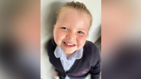 Arabella smiling at the camera. The image is taken from the top, looking down at the little girl. Her smiling face is in focus, with the rest of the image blurred. She has blonde hair tied back and blue eyes. She is wearing a blue and white blouse and blue blazer.