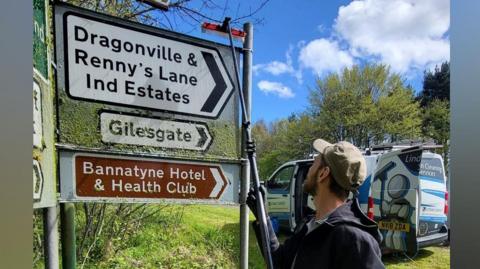 Jonny Linden has a cap on in the sunshine and is cleaning a mossy road sign with a brush. The sign reads 'Dragonville & Renny's Lane Ind Estates and Gilesgate. Beneath is a brown sign reading 'Bannatyne Hotel & Health Club".
