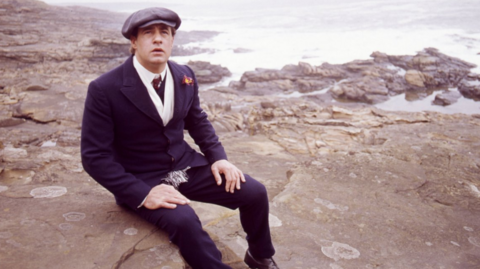 David appearing in Centre Play: God Bless Thee Jacky Maddison - Play by Tom Hadaway. Picture shows David Daker as Jacky Maddison. He is sat on rocks on a beach with a hat on.