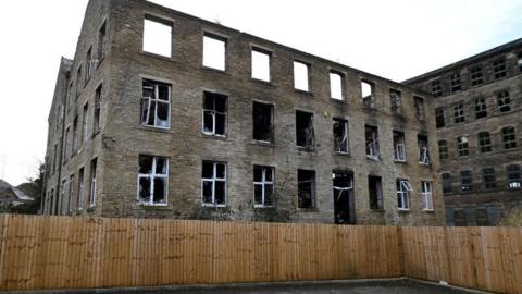A mill building which has been damaged by fire, the roof has gone as have many of the windows and there is a fence around which looks new. 