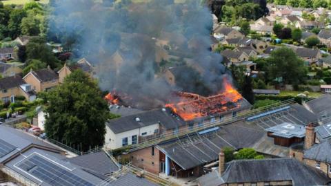 A large building is engulfed in flames, its roof collapsed and thick smoke rising into the sky