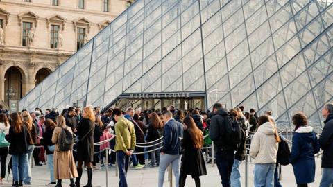 A winding queue of visitors outside the entrance to the Louvre, which is a tall glass pyramid intercut with a black criss-crossed pattern.