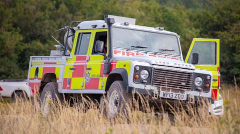 A silver fire service Land Rover in long dry grass. It has red and yellow checker pattern down the side.