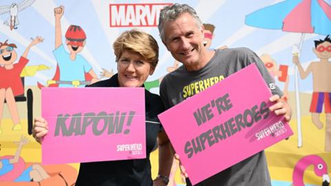 Clare Balding, a woman with short blonde hair, and Adam Hills a man with grey hair, hold up pink signs which read "kapow!" and "we're superheroes"