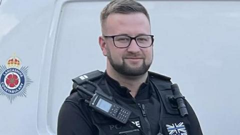 Head and shoulders of Alex Rose in full police uniform standing at the side of a white police van with the Lancashire Constabulary crest to the left. He has short dark hair, a short beard and black-framed glasses