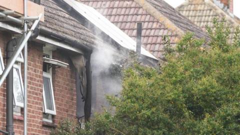 A charred building with smoke coming out of it. A green bush is in the foreground, shielding the house from view.