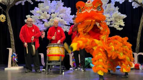 A bright orange lion puppet is dancing to drums and cymbols. 