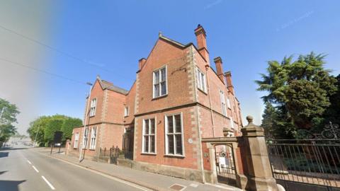 Bass House, a red brick building with ornate railing alongside it, fronts onto a main road. Behind the railings is a tree and in the distance to the left of the image there is a road junction.
